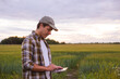 © Acronym - Farmer with a tablet computer in front of a sunset agricultural landscape. Countryside field. The concept of country life, food production, farming and technology concept.