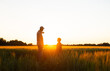 © Acronym - Farmer and his son in front of a sunset agricultural landscape. Man and a boy in a countryside field. Fatherhood, country life, farming and country lifestyle concept.