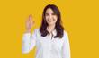 © Studio Romantic - Glad to see you. Happy, friendly woman says Hi. Studio shot of charming young brunette girl in white office shirt standing isolated on yellow background, waving hand, saying hello and smiling