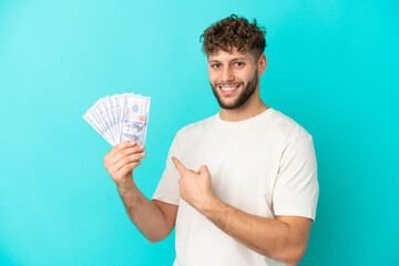 Canvas Print - Young caucasian man taking a lot of money isolated on blue background pointing back