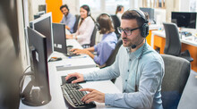 Male Customer Service Representative With Headset Working In Call Center Office With Colleagues In Row.