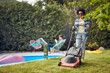 Woman Mowing the Lawn as Her Husband Relaxes by the Pool