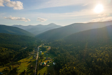 Wall Mural - Aerial view of amazing scenery with small village houses between foggy dark mountain peaks covered with forest pine trees at autumn sunrise. Beautiful wild woodland with shining rays of light at dawn