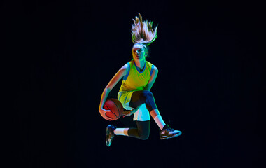 Wall Mural - Dynamic image of motivated sportsman, young girl playing basketball, jumping on black studio background in neon light. Concept of professional sport, action and motion, game, competition, hobby, ad