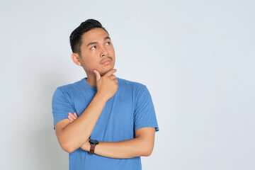 Pensive young Asian man in blue t-shirt looking at copy space, touching his chin, thinking and making decision isolated on white background