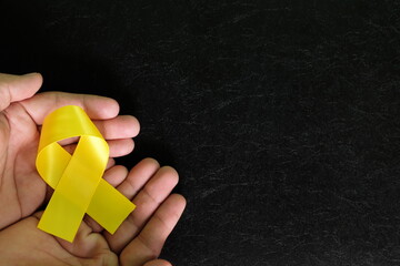 Human hands holding yellow ribbon color on dark black background. Bone cancer awareness and suicide prevention hope concept.