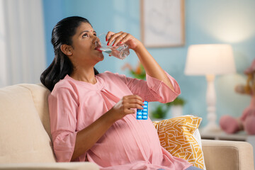 Indian pregnant woman taking pills or medicine while sitting on sofa at home - concept of Pregnancy wellness, Maternal medication and motherhood