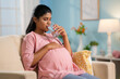© WESTOCK - Indian pregnant woman drinking water while sitting on sofa at home - concept of hydration, healthy pregnancy lifestyle and wellness