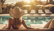 © annebel146 - Back view of young stylish woman with long blond hair wearing straw hat on her vacation at a beautiful resort, standing by a warm summer swimming pool with blue water on a sunny day.