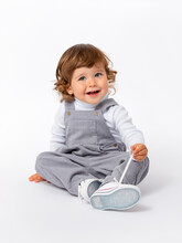 A Cheerful, Mischievous 2-year-old Toddler With Curly Hair In A Gray Jumpsuit And A White Turtleneck Sits On A White Background And Plays With Shoe Laces. Expressive Look.