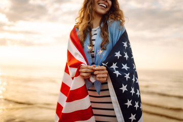 Young woman with American flag  and sparklers on the beach. Patriotic holiday. USA celebrate 4th of July. Independence Day concept