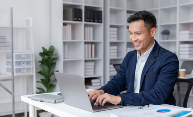 Confident Asian businessman working with laptop and papers on desk at office.