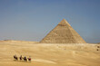 caravana de camellos con turistas por el desierto con una pirámide de fondo 