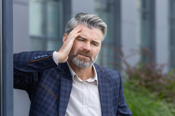 Wall Mural - Mature boss sick, man has severe headache, businessman in business suit adult overworked holding hands behind head, gray haired worker outside office building with headache.