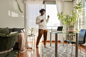 Happy funny gen z hipster African American teen guy wearing headphones dancing at home, listening music on mobile phone, having fun feeling funky moving in living room, authentic shot.