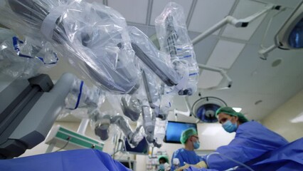 Wall Mural - Looking at the arms of the futuristic robot surgeon from low angle view. Medics stand at the patient in blur.