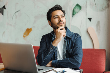 Handsome pensive Indian man, freelancer, manager using laptop computer working online sitting at workplace, planning startup. Asian student studying, learning languages, exam preparation, education