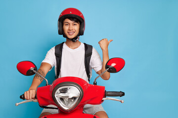 Wall Mural - Smiling oriental guy posing on his red motor bike, wearing red helmet, pointing at empty space aside, fast drive concept, copy space