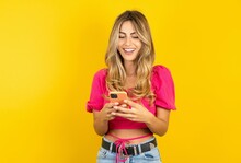 Young Blonde Woman Wearing Pink Crop Top Over Yellow Studio Background Using Mobile Phone Chatting Free Time .