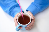 Fototapeta  - Kubek z gorącą herbatą trzymany w dłoniach, różowa plastikowa słomka jednorazowego użytku w środku 