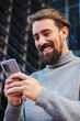 © Jose Calsina - Vertical portrait of happy business young male enjoying using a mobile phone app sharing media on internet. Awesome handsome man texting and laughing with cellphone outdoors. Guy using a mobile phone