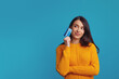 Thoughtful young girl wearing yellow clothes holding plastic credit card near face and looking at free empty space, isolated over blue background 