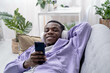 © Наталия Кузина - Black young man using smartphone at home lying on couch, pressing finger, reading social media internet, typing text or shopping online, Making video call
