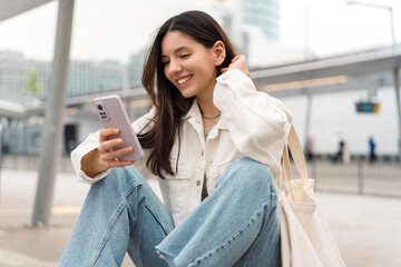 Bright portrait of cheerful young woman 25s with shopper on shoulder waiting public transport at station smiling beautifully and texting