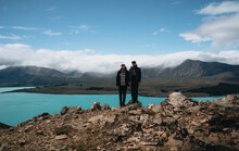 Two Young Male Tourist Stand In Front Of Blue Lake Tekapo In New Zealand, Canterbury Region. Front View Of Young Man In Hat Standing On Small Hill At Mount John Observatory.