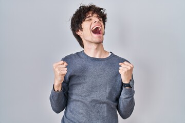 Poster - Young man standing over isolated background celebrating surprised and amazed for success with arms raised and eyes closed. winner concept.