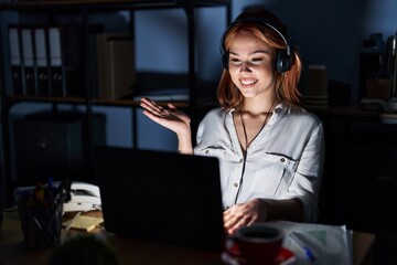 Poster - Young caucasian woman working at the office at night smiling cheerful presenting and pointing with palm of hand looking at the camera.