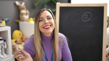 Sticker - Young blonde woman preschool teacher smiling confident holding chalk by blackboard at kindergarten