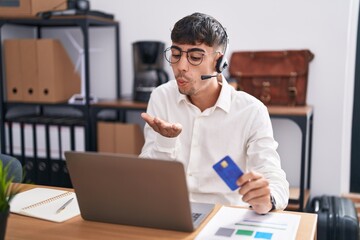 Wall Mural - Young hispanic man working using computer laptop holding credit card looking at the camera blowing a kiss with hand on air being lovely and sexy. love expression.