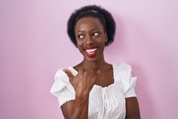 Wall Mural - African woman with curly hair standing over pink background smiling with happy face looking and pointing to the side with thumb up.