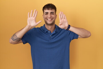 Wall Mural - Young hispanic man standing over yellow background showing and pointing up with fingers number nine while smiling confident and happy.