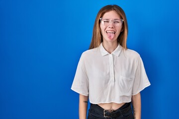 Poster - Beautiful woman standing over blue background sticking tongue out happy with funny expression. emotion concept.