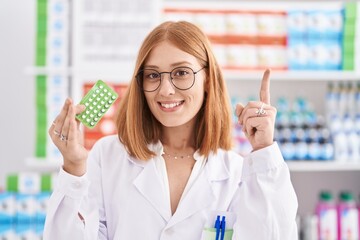 Wall Mural - Young redhead woman working at pharmacy drugstore holding birth control pills surprised with an idea or question pointing finger with happy face, number one