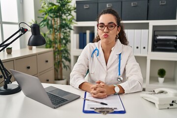 Canvas Print - Young hispanic woman wearing doctor uniform and stethoscope puffing cheeks with funny face. mouth inflated with air, crazy expression.