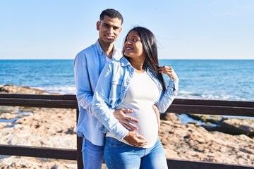 Wall Mural - Young latin couple expecting baby hugging each other standing at seaside