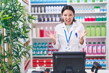 Sticker - Hispanic young woman working at pharmacy drugstore crazy and mad shouting and yelling with aggressive expression and arms raised. frustration concept.