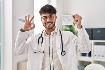 Canvas Print - Hispanic doctor man with beard holding covid record card doing ok sign with fingers, smiling friendly gesturing excellent symbol