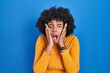Black woman with curly hair standing over blue background afraid and shocked, surprise and amazed expression with hands on face
