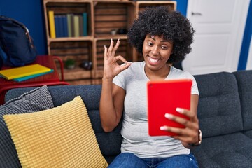 Sticker - Black woman with curly hair using touchpad sitting on the sofa doing ok sign with fingers, smiling friendly gesturing excellent symbol