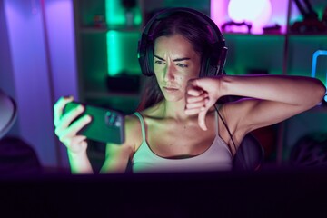 Poster - Young brunette woman playing video games with smartphone with angry face, negative sign showing dislike with thumbs down, rejection concept