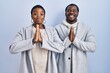 Young african american couple standing over blue background together praying with hands together asking for forgiveness smiling confident.
