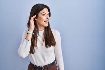 Sticker - Young brunette woman standing over blue background smiling with hand over ear listening an hearing to rumor or gossip. deafness concept.