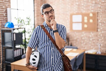 Wall Mural - Hispanic man with long hair working at the office holding bike helmet with hand on chin thinking about question, pensive expression. smiling with thoughtful face. doubt concept.