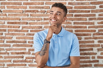 Sticker - Brazilian young man standing over brick wall with hand on chin thinking about question, pensive expression. smiling and thoughtful face. doubt concept.