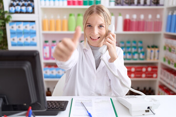 Poster - Young caucasian woman working at pharmacy drugstore speaking on the telephone approving doing positive gesture with hand, thumbs up smiling and happy for success. winner gesture.