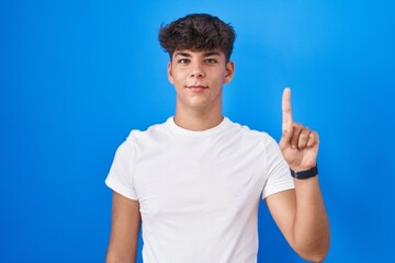 Canvas Print - Hispanic teenager standing over blue background showing and pointing up with finger number one while smiling confident and happy.